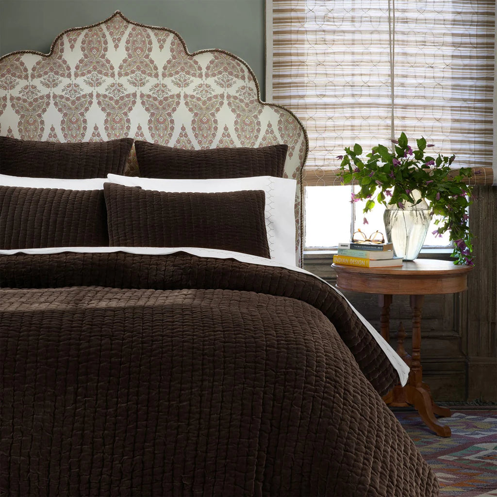 A cozy bedroom features a decorative upholstered headboard, John Robshaw’s Velvet Coverlet King in dark brown with matching pillows, and a round wood bedside table topped with a glass vase of green foliage and stacked books by the window.