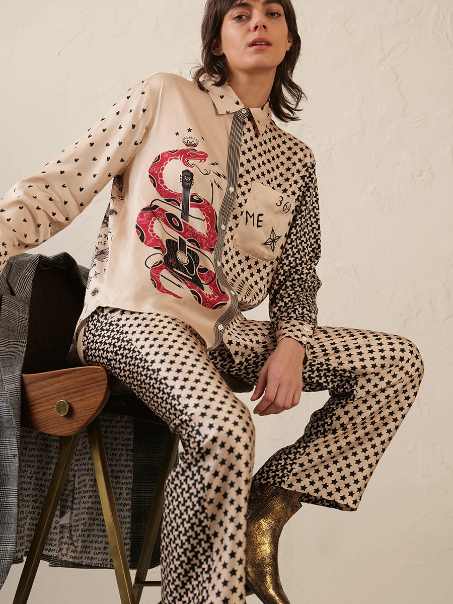 A person sits on a wooden chair wearing Me369 Marilyn Trousers—cream-colored straight leg pants with black stars and a bold red snake graphic—paired with gold ankle boots, against a light textured wall.