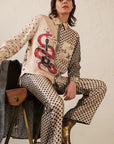 A person sits on a wooden chair wearing Me369 Marilyn Trousers—cream-colored straight leg pants with black stars and a bold red snake graphic—paired with gold ankle boots, against a light textured wall.