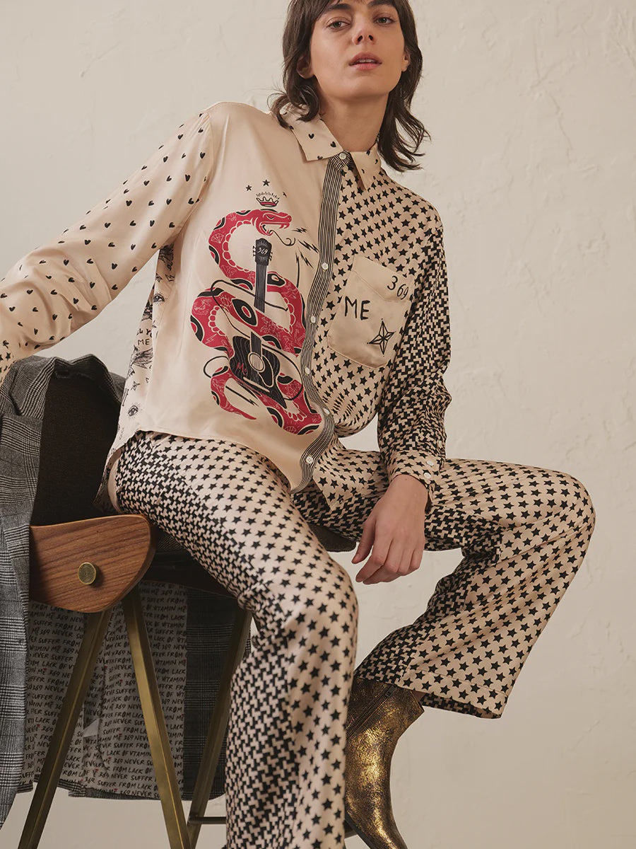 A person sits on a chair wearing the Me369 Scarlett Print Shirt with black stars, red snake, and guitar motifs, paired with matching star pants and shiny gold ankle boots.