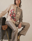 A person sits on a chair wearing the Me369 Scarlett Print Shirt with black stars, red snake, and guitar motifs, paired with matching star pants and shiny gold ankle boots.
