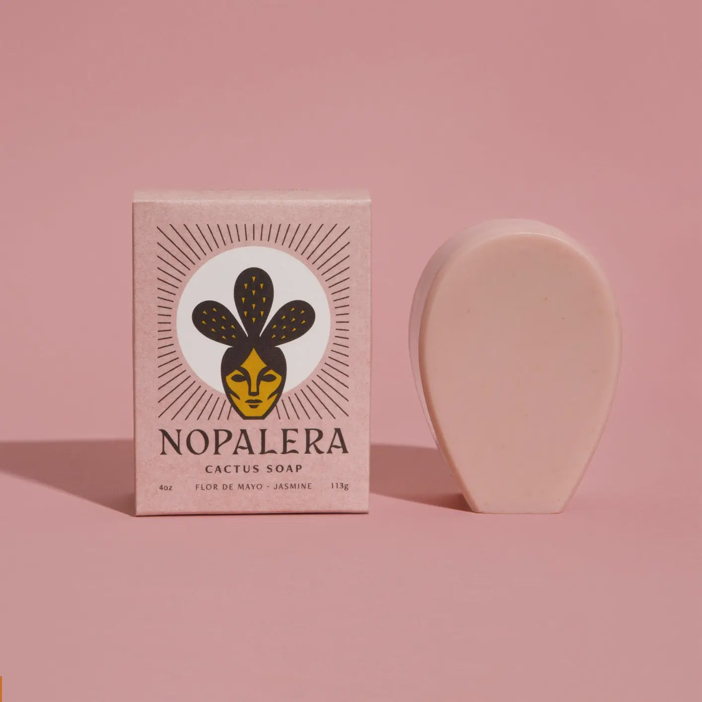 A pink box of Flor De Mayo Jasmine Soap by Faire, enriched with Pink Rose Clay, sits against a pink backdrop. The cactus pad-shaped soap mirrors the artistic box design, showcasing a face and plant theme that captures the essence of Prickly Pear Cactus.