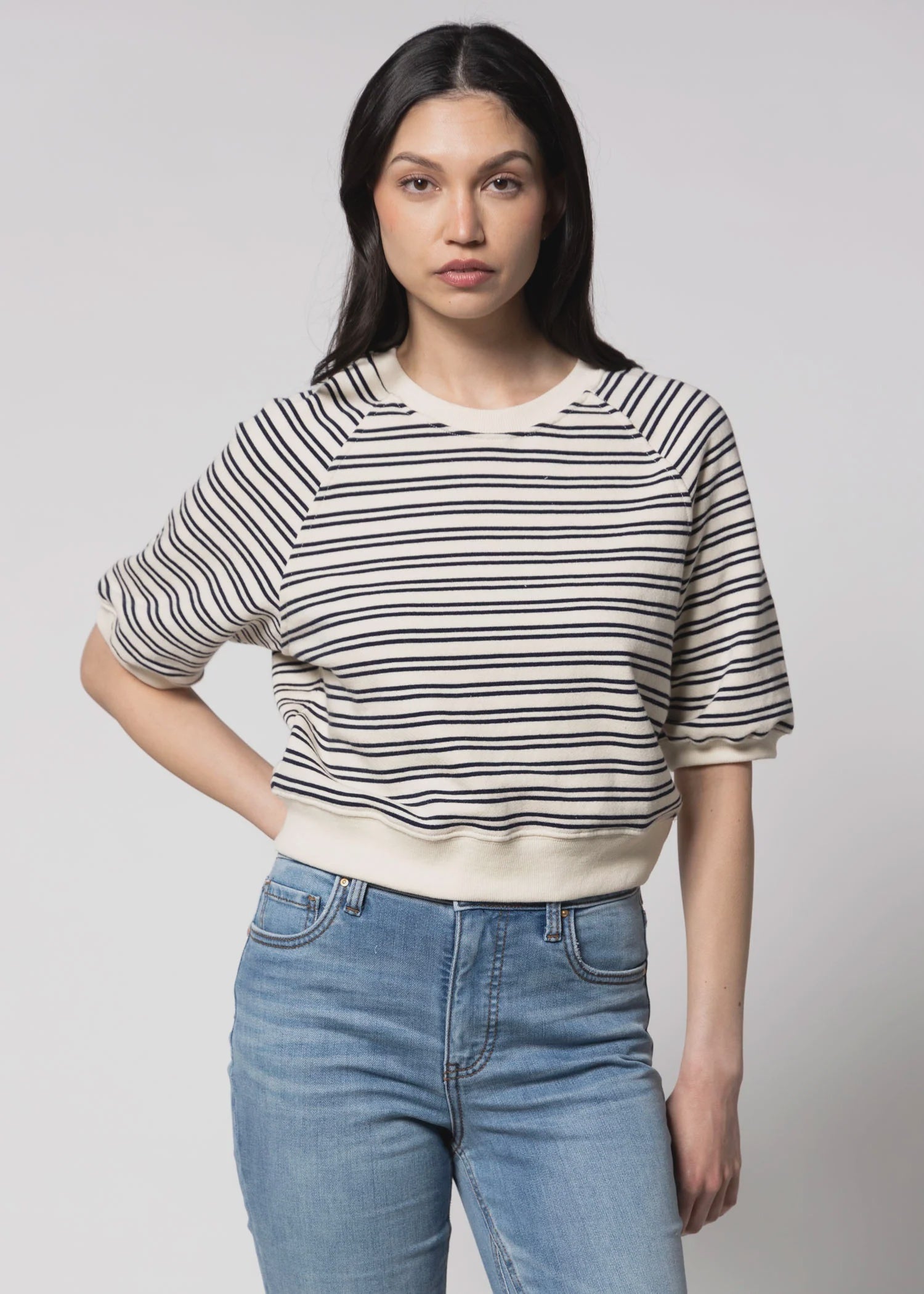 A woman with long dark hair wears the Perfectwhitetee French Terry SS Raglan Sweatshirt, a 100% cotton cream short-sleeve top with black stripes, paired with high-waisted light blue jeans. She stands against a plain light background, hand on hip.