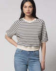 A woman with long dark hair wears the Perfectwhitetee French Terry SS Raglan Sweatshirt, a 100% cotton cream short-sleeve top with black stripes, paired with high-waisted light blue jeans. She stands against a plain light background, hand on hip.