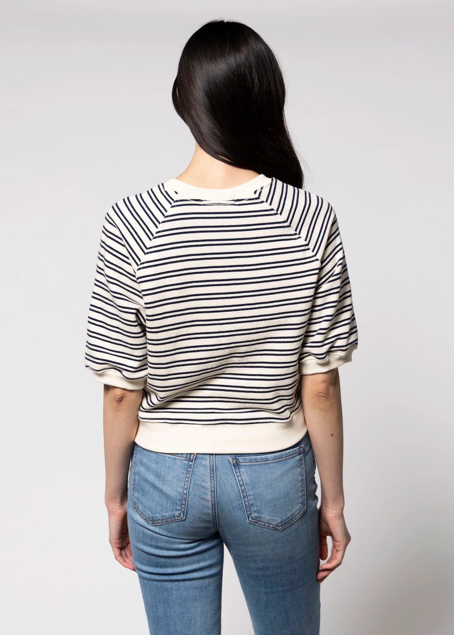 A person with long dark hair stands with their back to the camera against a plain light background, wearing the Perfectwhitetee French Terry SS Raglan Sweatshirt in white and navy stripes with blue jeans.