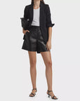 A person dons a black blazer over a white top paired with Frame's chic Leather Trouser Shorts, featuring a mid-rise waistband and side slash pockets. Completing the look are black heeled sandals and a sleek black clutch against a plain backdrop.