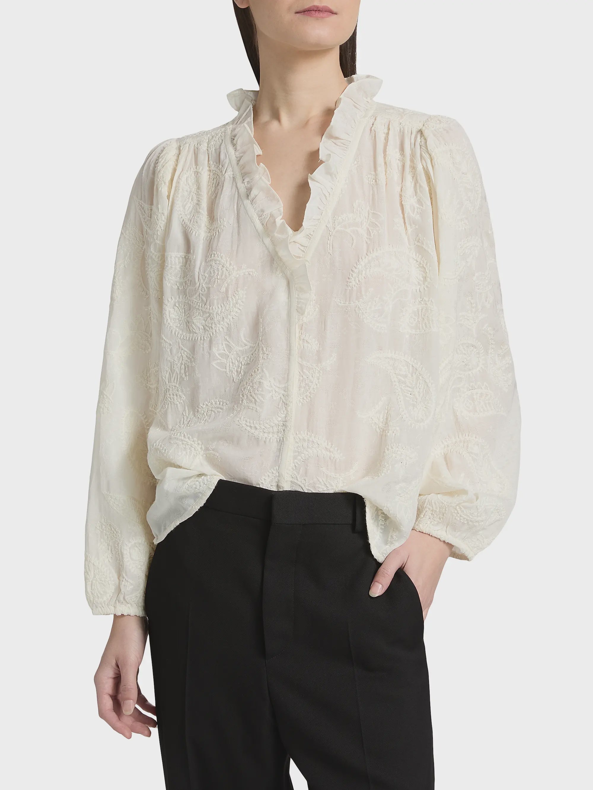 A woman wears the BA&SH Shirt Petty—a long-sleeved, cream blouse with ruffles and paisley embroidery—paired with black trousers. She stands with one hand in her pocket against a plain background.