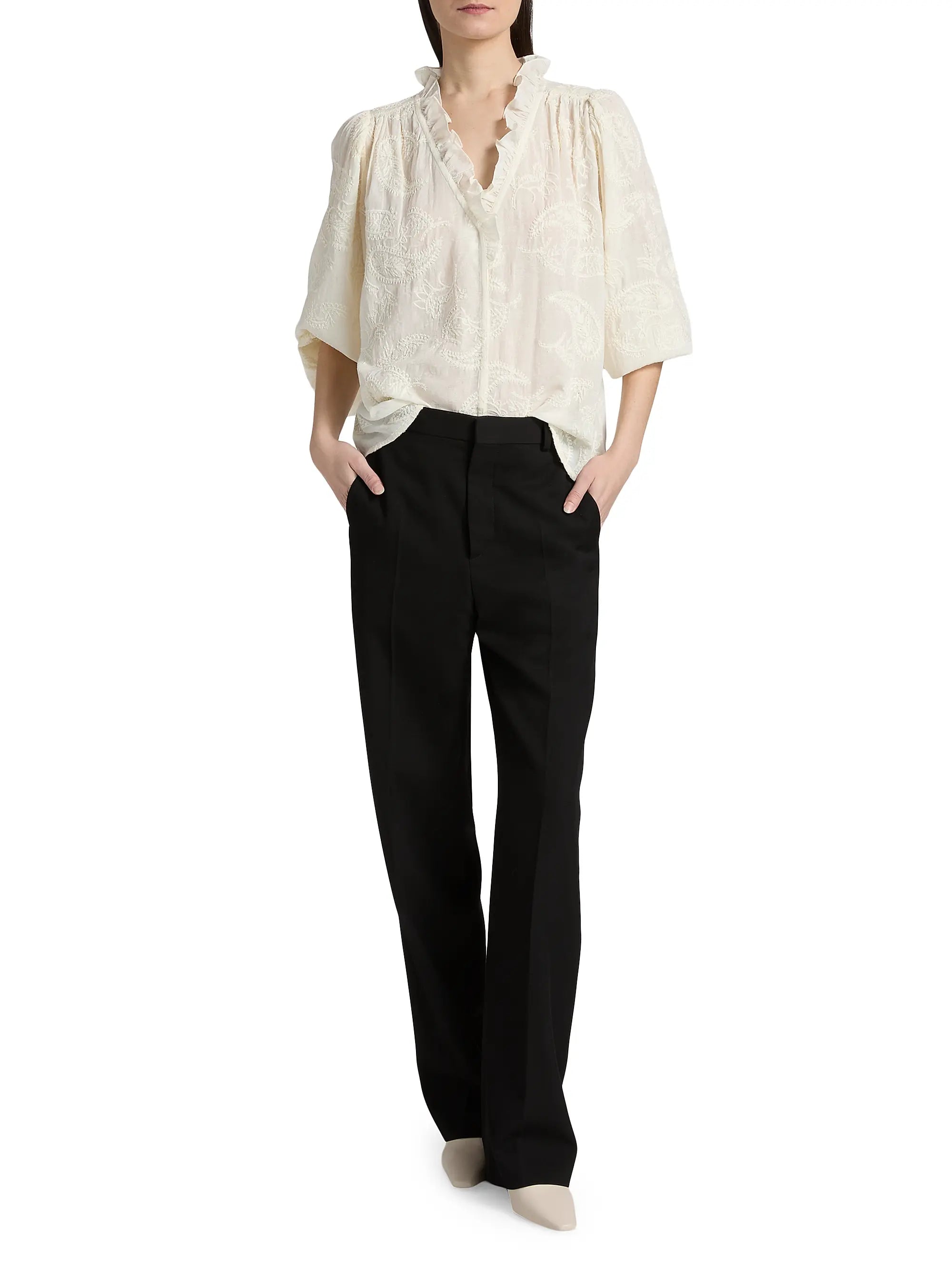 A person stands with hands in pockets, wearing the BA&SH Shirt Petty—a loose, cream-colored bohemian blouse with short sleeves—paired with black straight-leg trousers and light-colored shoes.