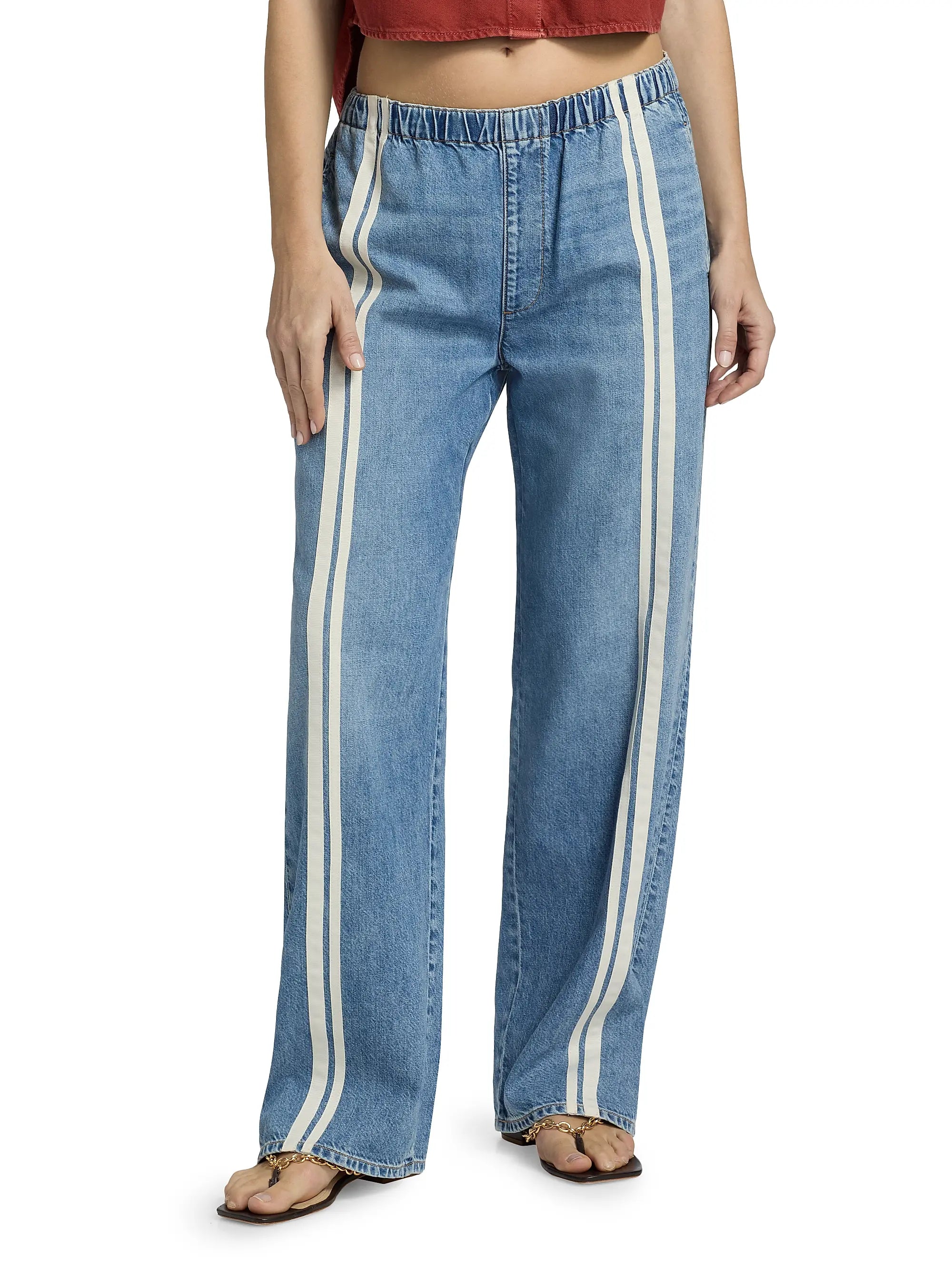 A person is seen from the waist down wearing Rag & Bone ftwt logan pull on track jeans in blue with vertical white side stripes, paired with a rust cropped top and beige sandals.
