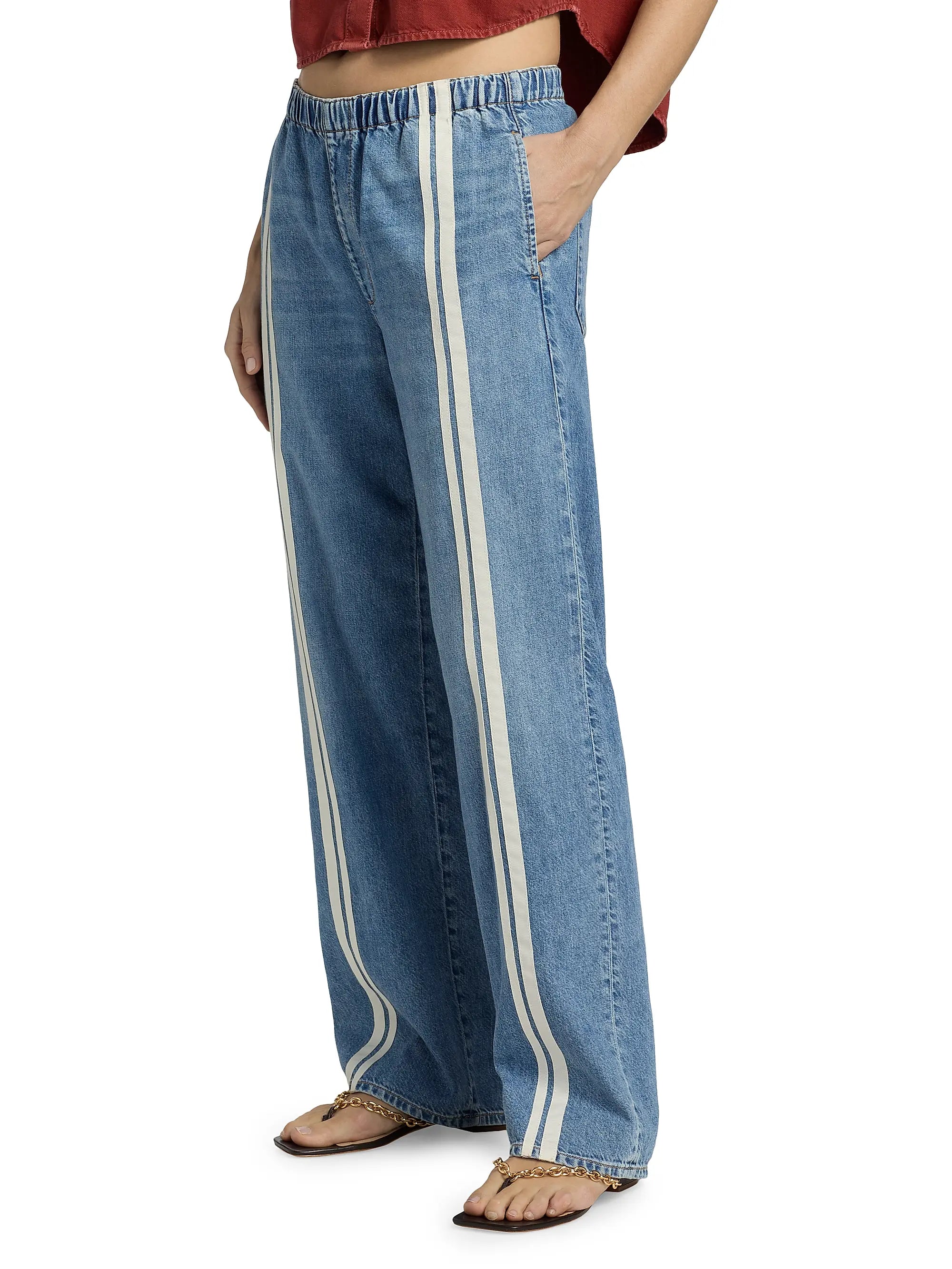A person wears Rag & Bone ftwt logan pull on track jeans—relaxed straight fit, cotton-blend denim with two vertical white stripes, elastic waistband—paired with a rust crop top and sandals. Only their lower half is visible, hands in pockets.
