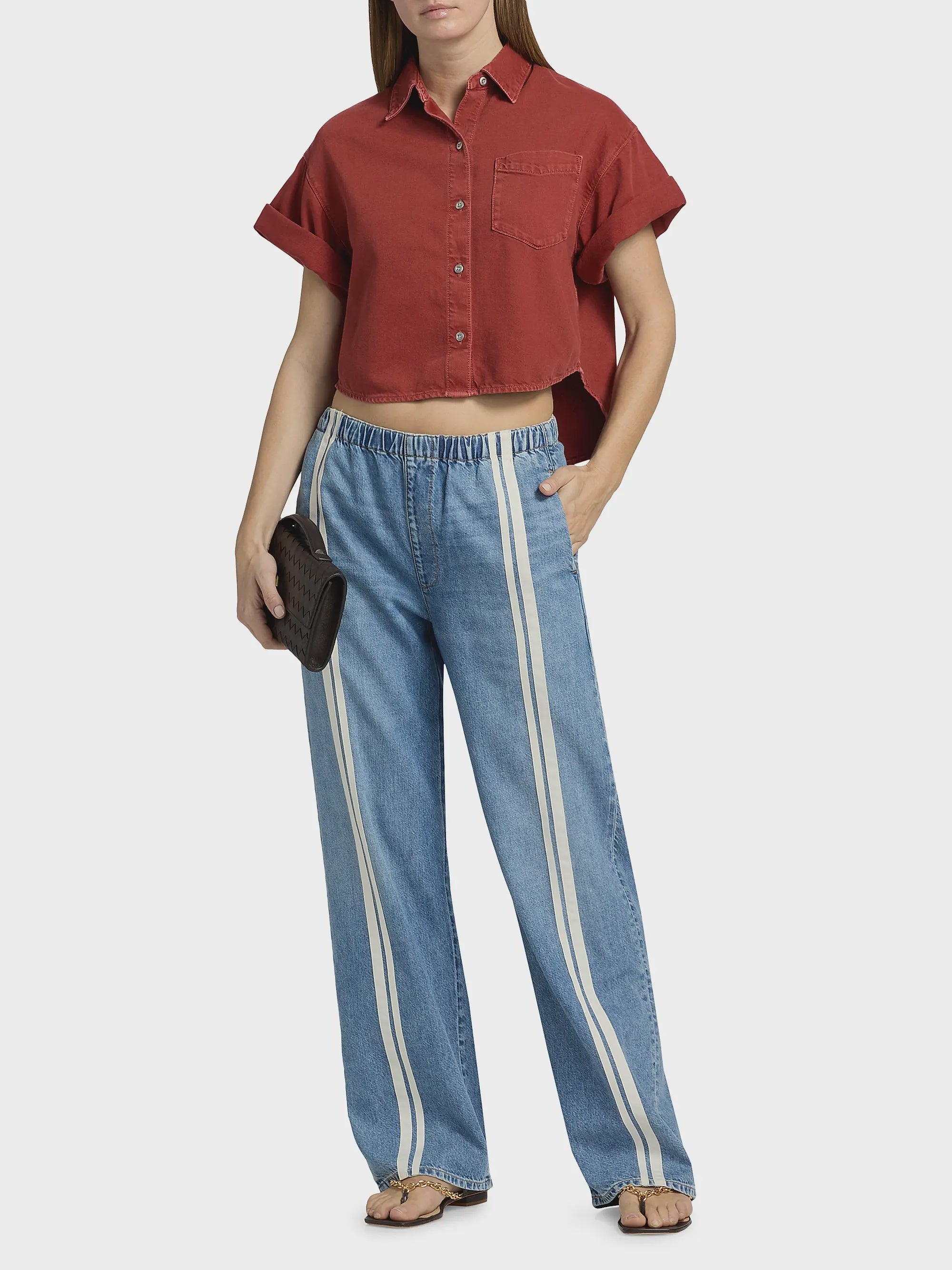 A woman wears a short-sleeve red cropped button-up shirt, Rag & Bone ftwt logan pull on track featherweight denim relaxed straight fit jeans with white side stripes, brown sandals, and holds a small dark clutch. One hand is in her pocket.