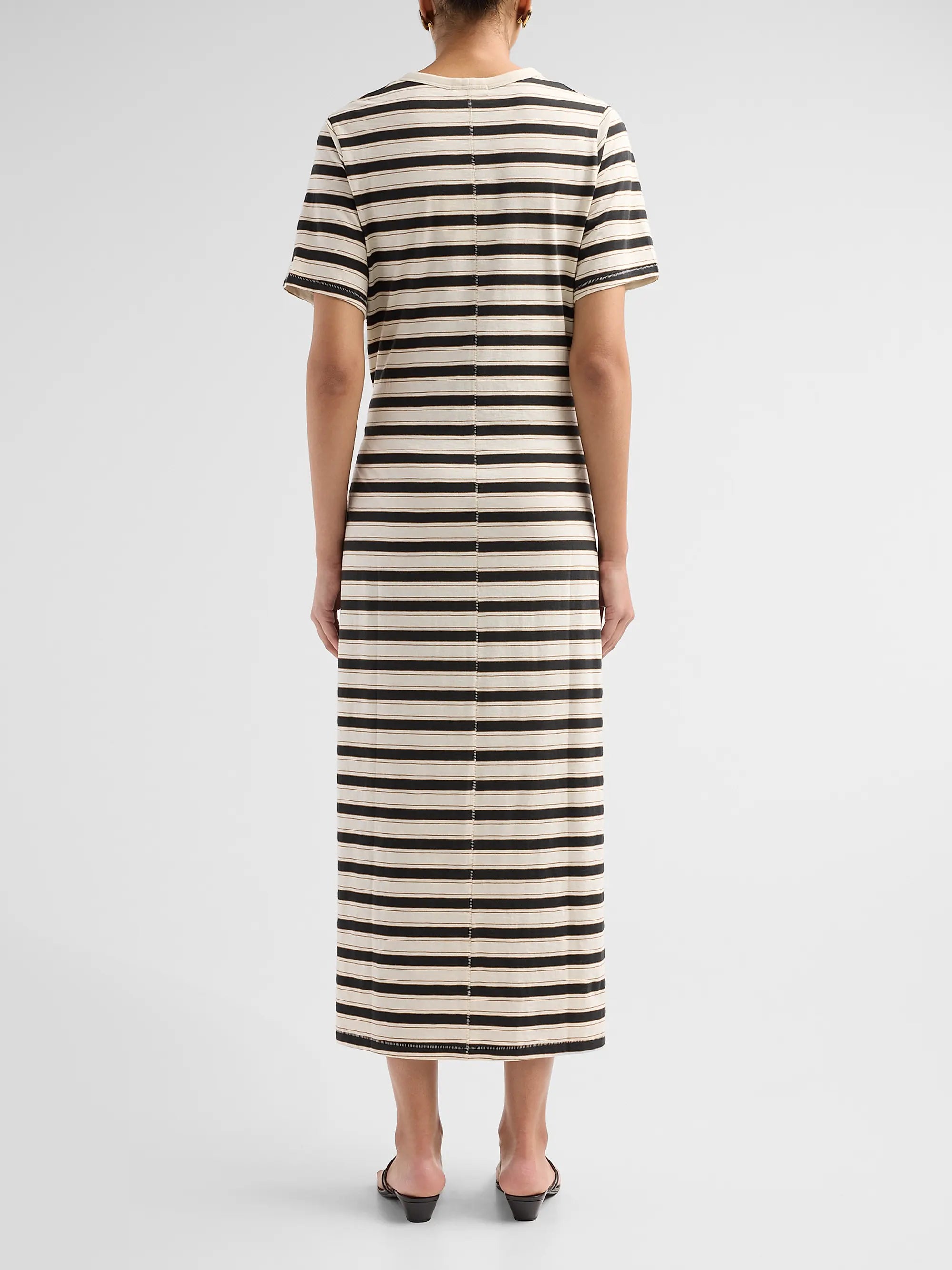 A person faces away, wearing Rag &amp; Bone's the slub stripe tee midi dress—a short-sleeved, ankle-length cotton jersey with black, white, and beige stripes—paired with black heeled shoes for a classic look.