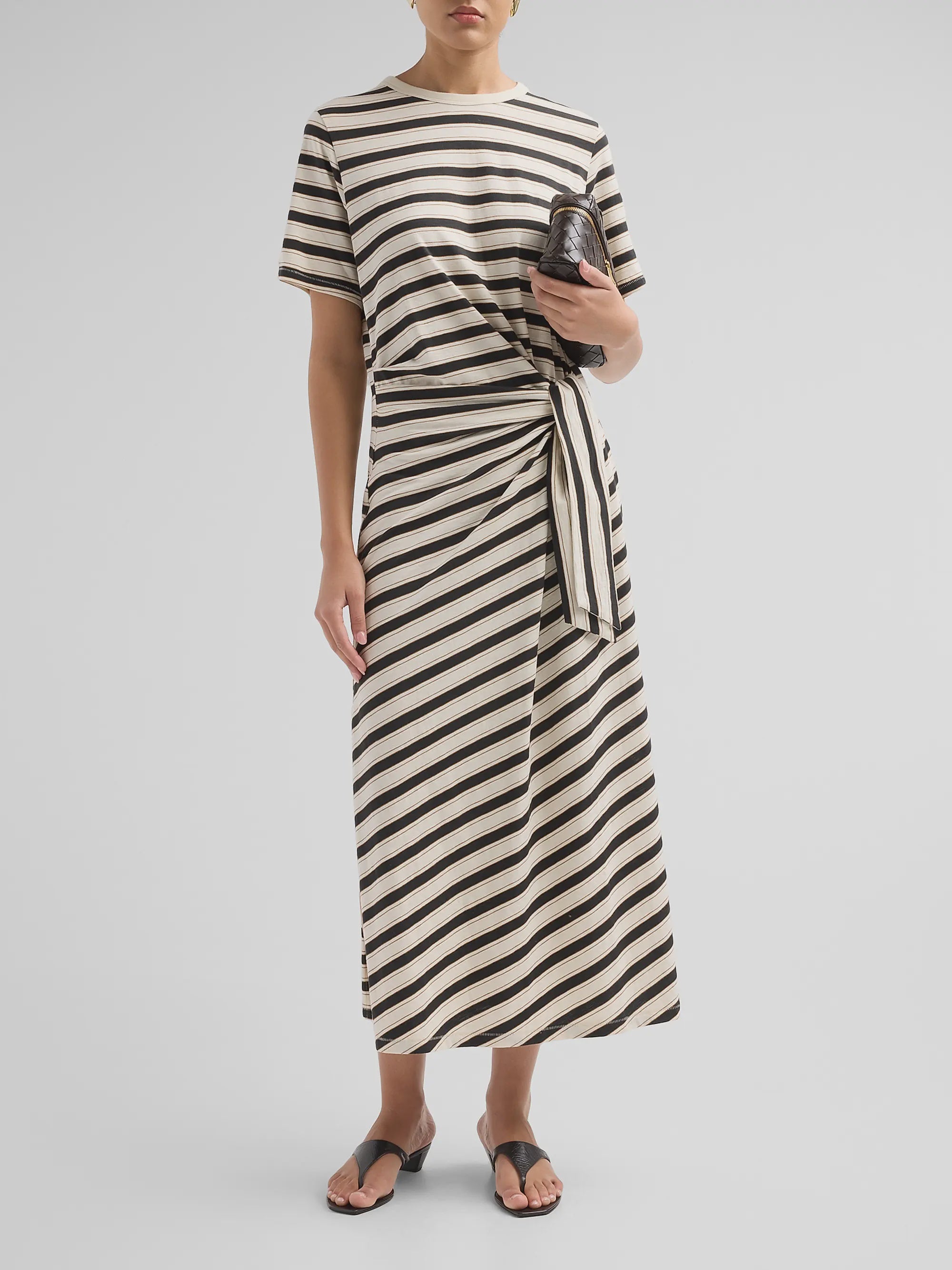 A person stands against a plain light background wearing the slub stripe tee midi dress by Rag &amp; Bone, styled with a knotted waist, holding a small clutch bag and wearing black sandals.