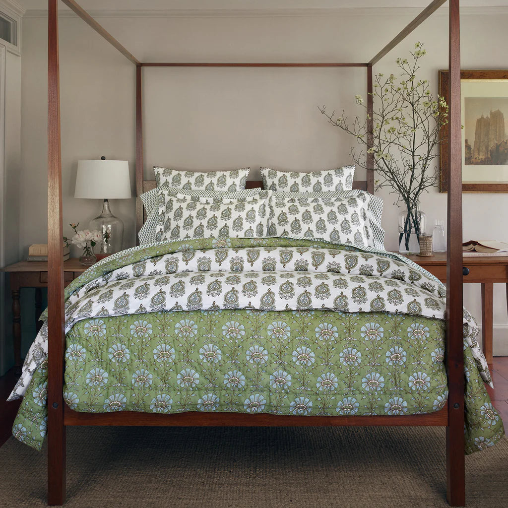 A cozy bedroom centers on a wooden four-poster bed dressed in the John Robshaw Jeeva Sage Quilt and white floral bedding, with neatly arranged pillows. Behind the bed, a desk holds a lamp, vase of branches, and framed art.