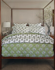 A cozy bedroom centers on a wooden four-poster bed dressed in the John Robshaw Jeeva Sage Quilt and white floral bedding, with neatly arranged pillows. Behind the bed, a desk holds a lamp, vase of branches, and framed art.