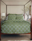 A cozy bedroom features a wooden four-poster bed adorned with the John Robshaw Jeeva Sage Quilt, floral bedding, light gray nightstands, a white table lamp, a vase with branches, and a framed picture on the wall.