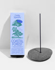 A box of Faire Low Smoke Incense, made with natural ingredients, sits beside a burning incense stick in a smooth round stone holder. The pale blue box shows green illustrations on a white background.