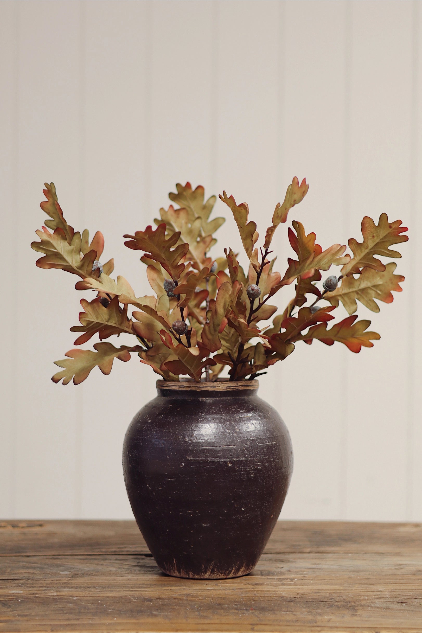 Faire Oak Leaf Spray in a dark brown ceramic vase by Bungalow Furniture, displayed on wood against a paneled wall.
