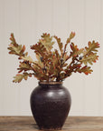 Faire Oak Leaf Spray in a dark brown ceramic vase by Bungalow Furniture, displayed on wood against a paneled wall.