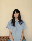 A person with long dark hair and bangs, wearing The Western Shirt by Hey Gang in an oversized light blue short-sleeve style, sits on a wooden bench against a beige wall, gazing to the side with a slight smile.