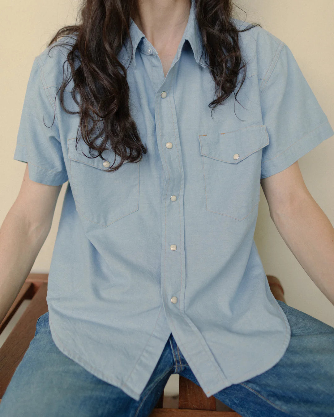 A person with long, dark hair is wearing The Western Shirt by Hey Gang, an oversized light blue top paired with blue jeans. They are sitting down with their arms resting on their knees against a plain and neutral background.