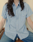 A person with long, dark hair is wearing The Western Shirt by Hey Gang, an oversized light blue top paired with blue jeans. They are sitting down with their arms resting on their knees against a plain and neutral background.