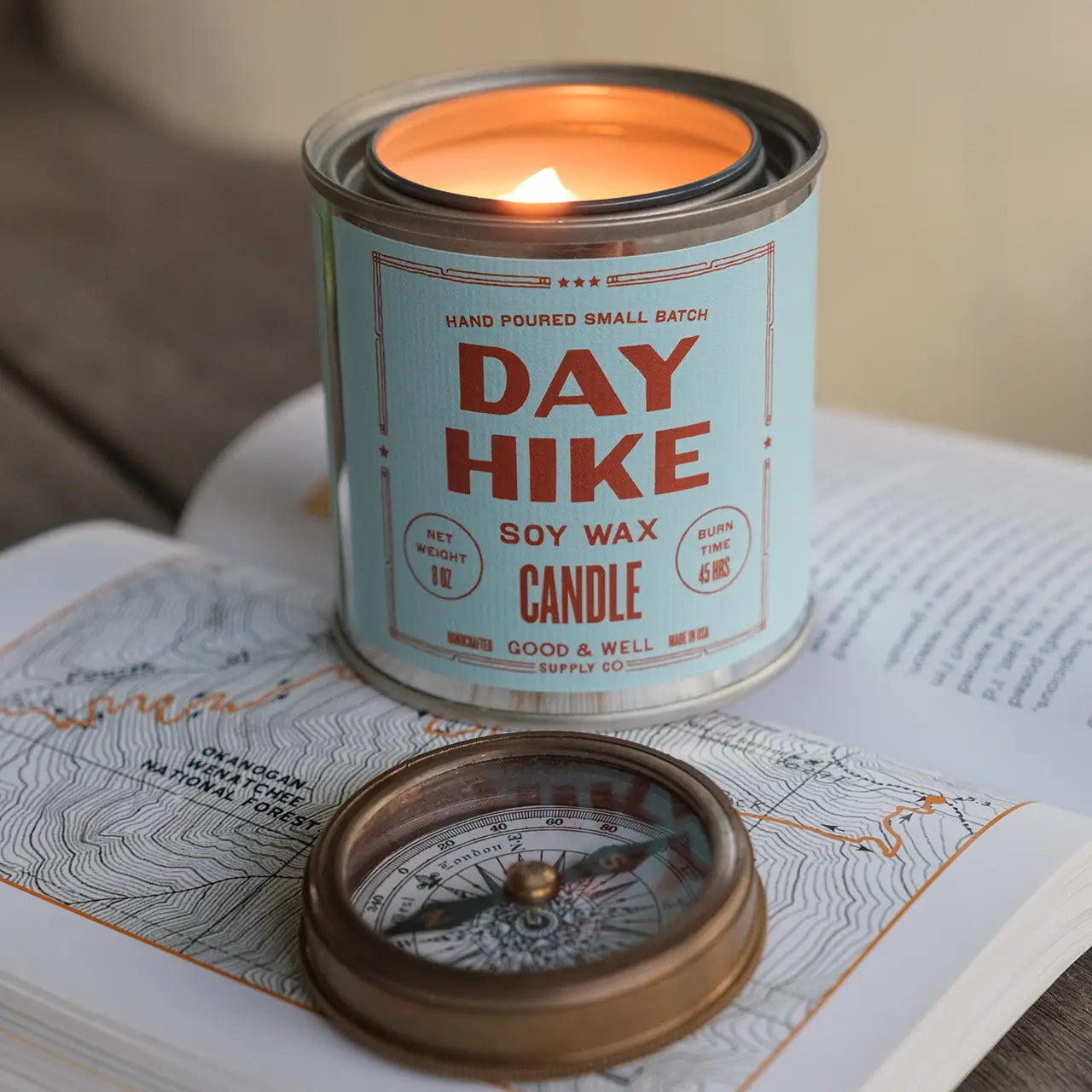 A lit G&W Soy Candle by Faire from the Happy Place Collection rests on an open book with a map beside a vintage compass, evoking nostalgic joy and inspiring thoughts of adventure and exploration.