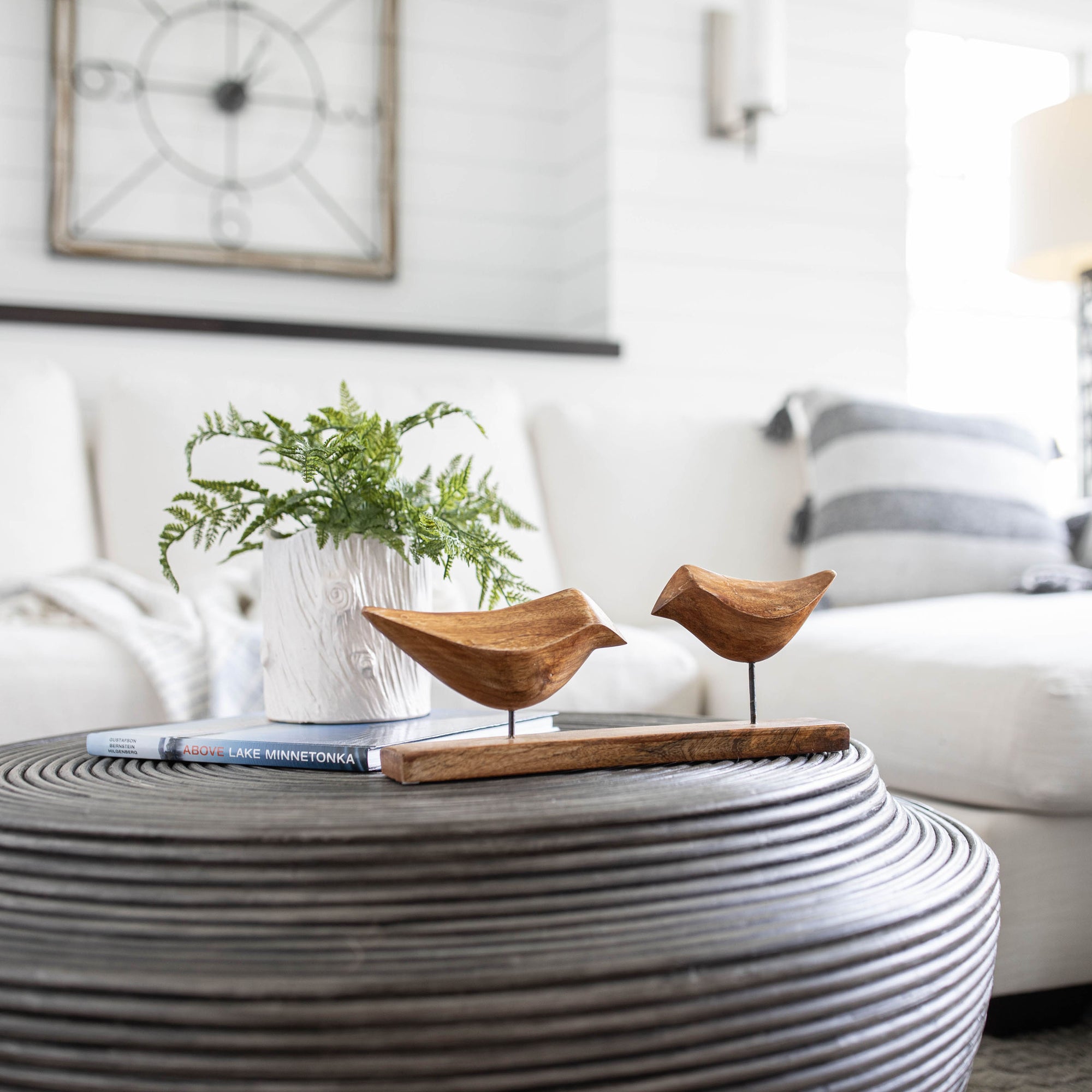 A round ribbed coffee table displays two Faire Arlo Natural Wood Decorative Birds, a white vase with green fern leaves, and a book in a bright, modern living room with a white sofa and striped pillow in the background.