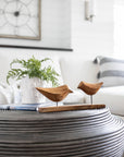 A round ribbed coffee table displays two Faire Arlo Natural Wood Decorative Birds, a white vase with green fern leaves, and a book in a bright, modern living room with a white sofa and striped pillow in the background.