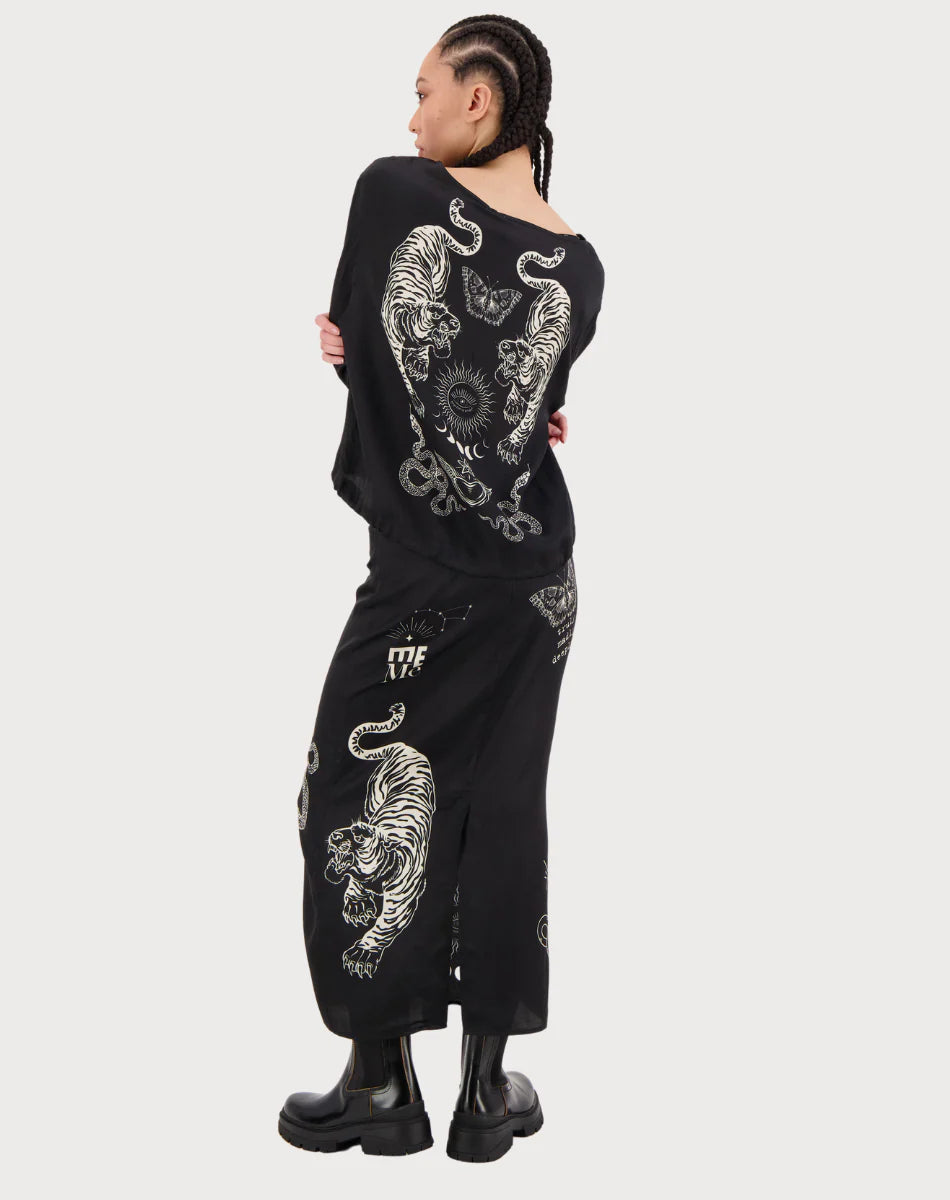 A person with braided hair stands facing away, wearing the Me369 Beatrice Blouson Top with a black magic print and white tiger and sun designs, paired with a matching skirt and black chunky boots.