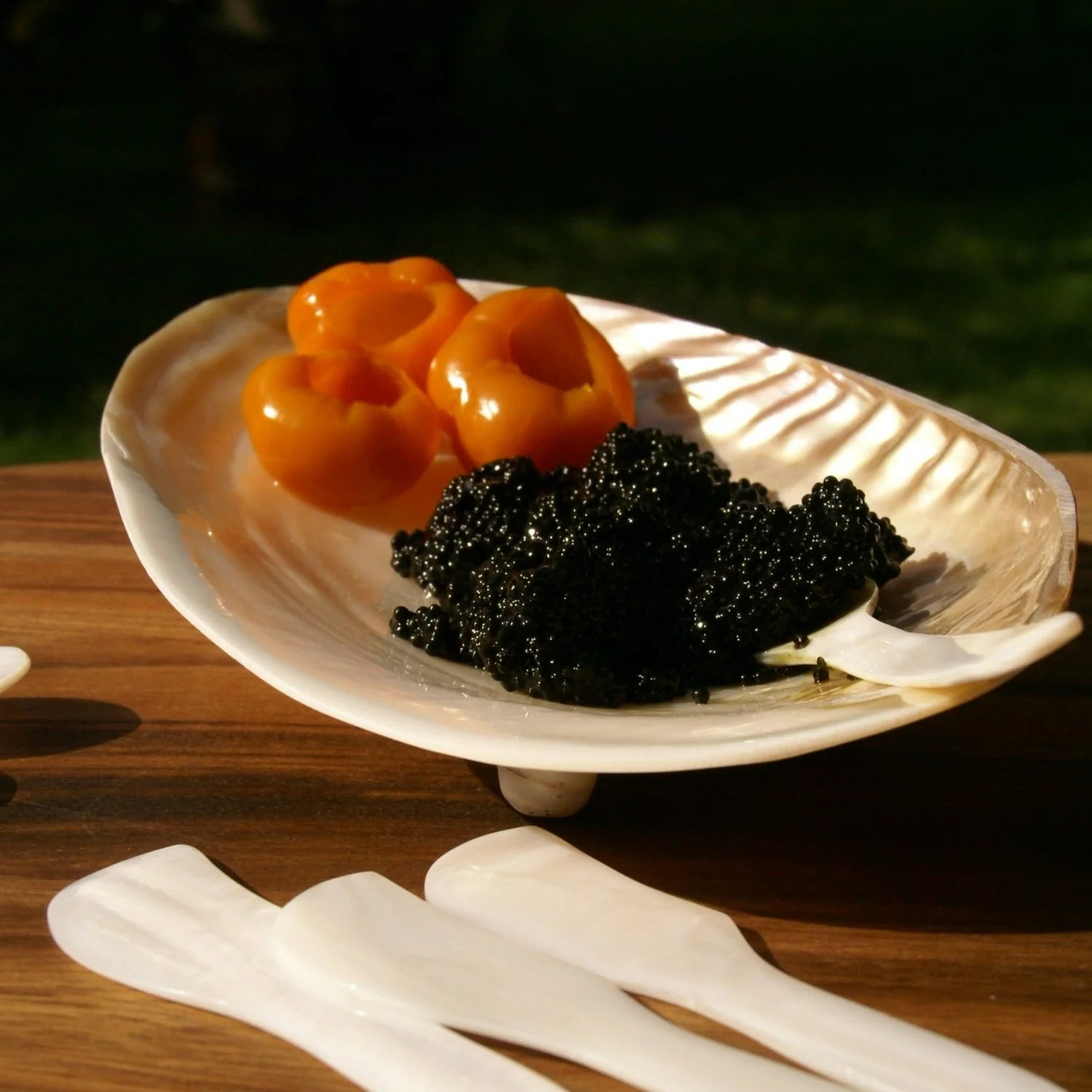 The Be Home 07-01 Seashell Footed Dish displays three golden yellow tomatoes and black caviar, set on wood with three white utensils—an ideal accent for lovers of ethically crafted nautical decor.