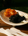 The Be Home 07-01 Seashell Footed Dish displays three golden yellow tomatoes and black caviar, set on wood with three white utensils—an ideal accent for lovers of ethically crafted nautical decor.