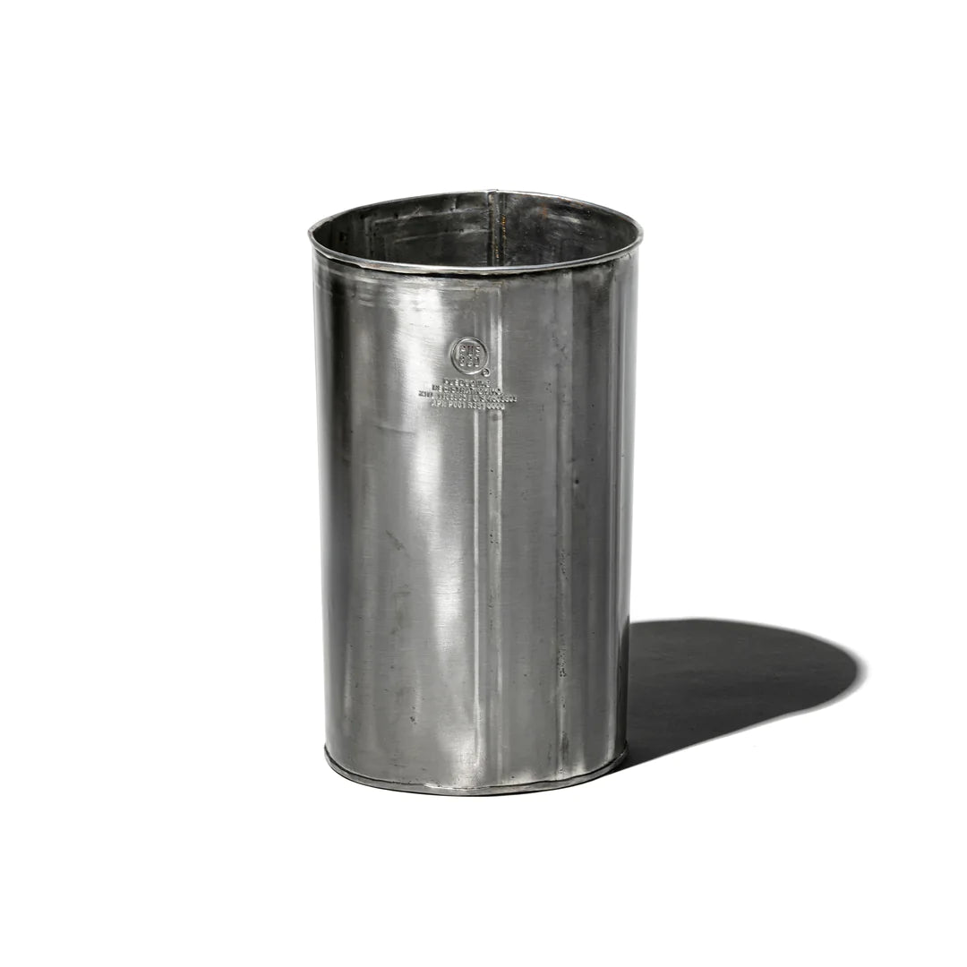 The Faire Steel Trashcan, a shiny cylindrical container made from recycled steel, stands upright on a white background. Its reflective surface casts a shadow to the right and displays a faint logo on the front.