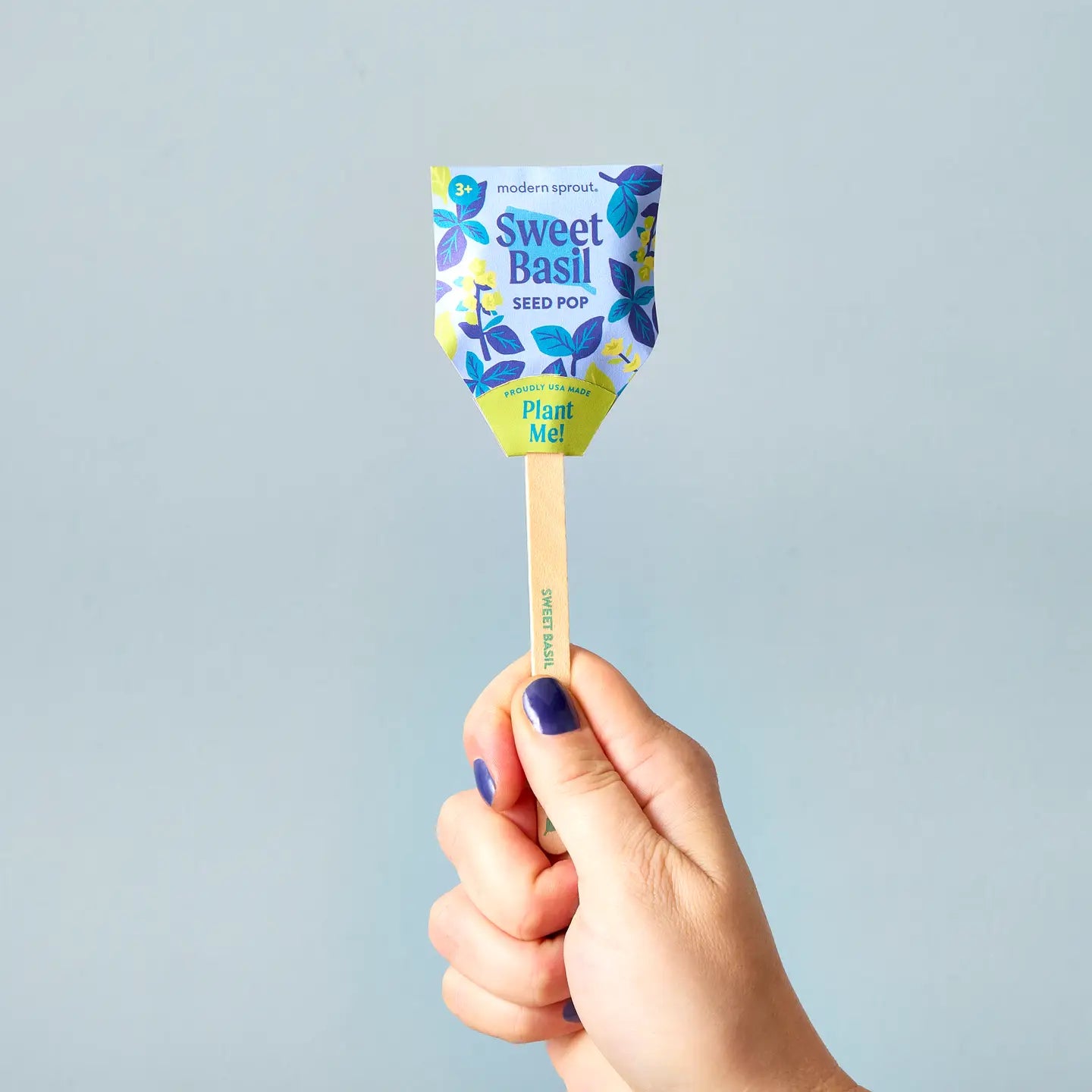 A hand with blue nail polish holds a Faire Garden Seed Pop on a wooden stick against a light blue background. The colorful packaging, ideal for seed starting, features leaves and text that says "Plant Me!".