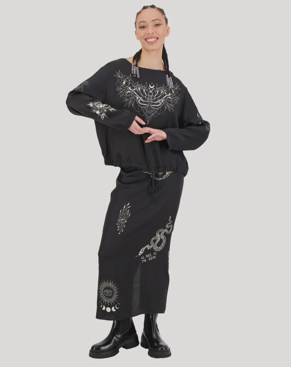 A woman with braided hair smiles, wearing a loose black top and the Me369 Valery Slim Skirt—a black midi with white mystical and celestial designs—paired with black boots. She stands against a plain light gray background.