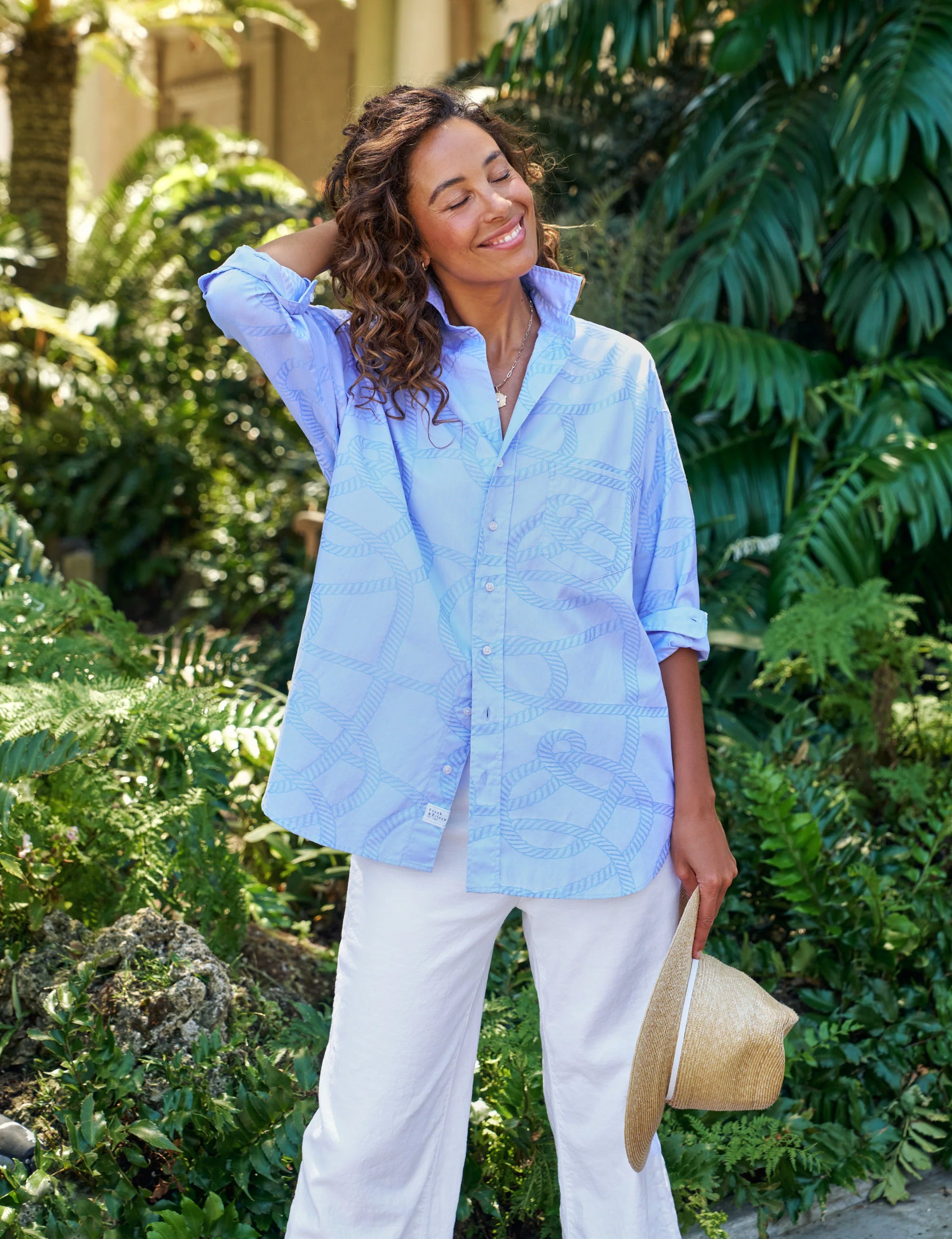 A smiling woman stands outdoors among green plants, wearing a relaxed-fit Frank & Eileen Eileen S25 light blue Italian Cotton shirt and white pants. She holds a straw hat and has her other hand behind her head, looking happy and relaxed.