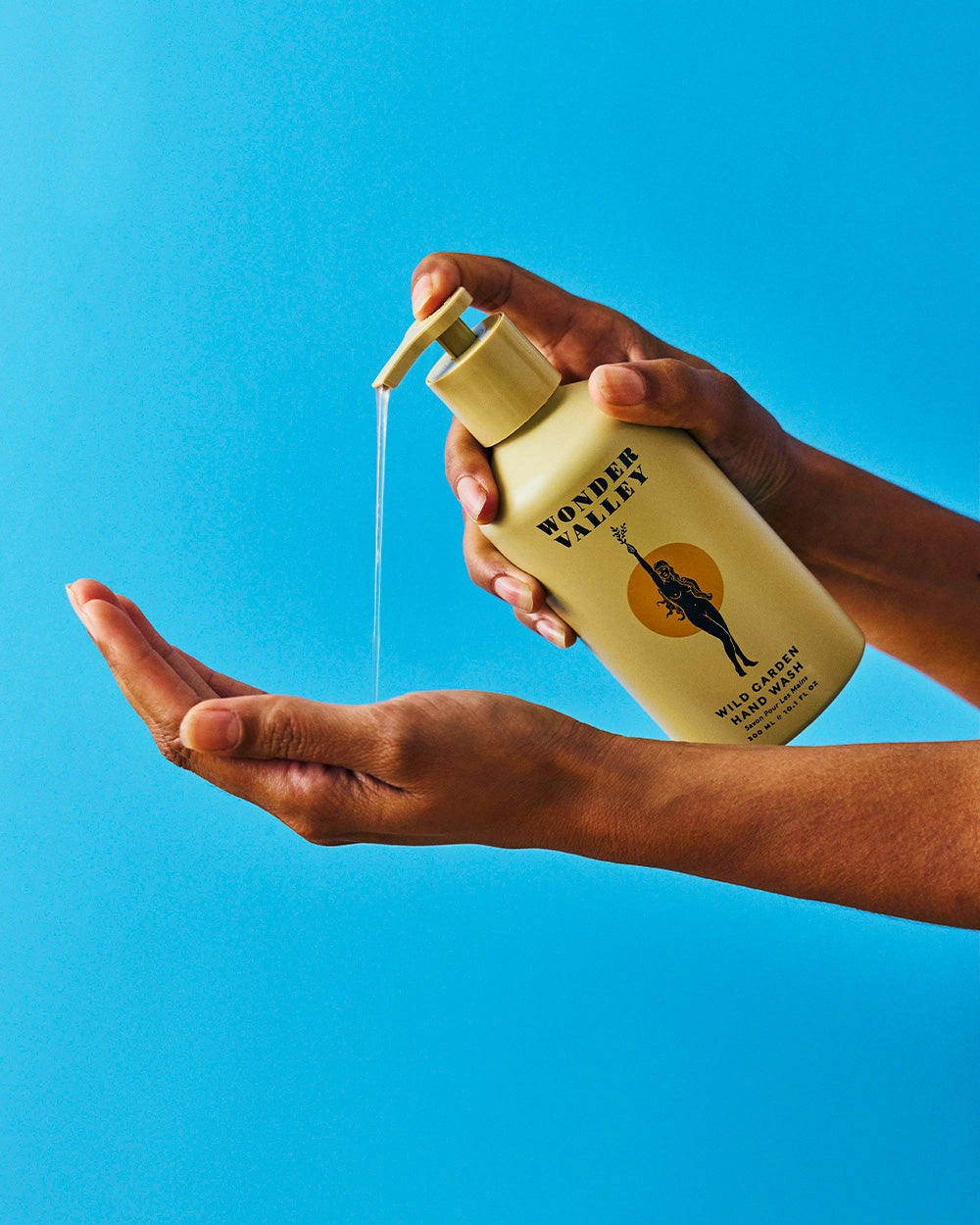 A person dispenses clear liquid from a yellow bottle labeled "Faire Wild Garden Hand Wash" into their palm, offering a deep clean and fresh herbal scent against a vibrant blue background.