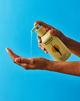 A person dispenses clear liquid from a yellow bottle labeled "Faire Wild Garden Hand Wash" into their palm, offering a deep clean and fresh herbal scent against a vibrant blue background.