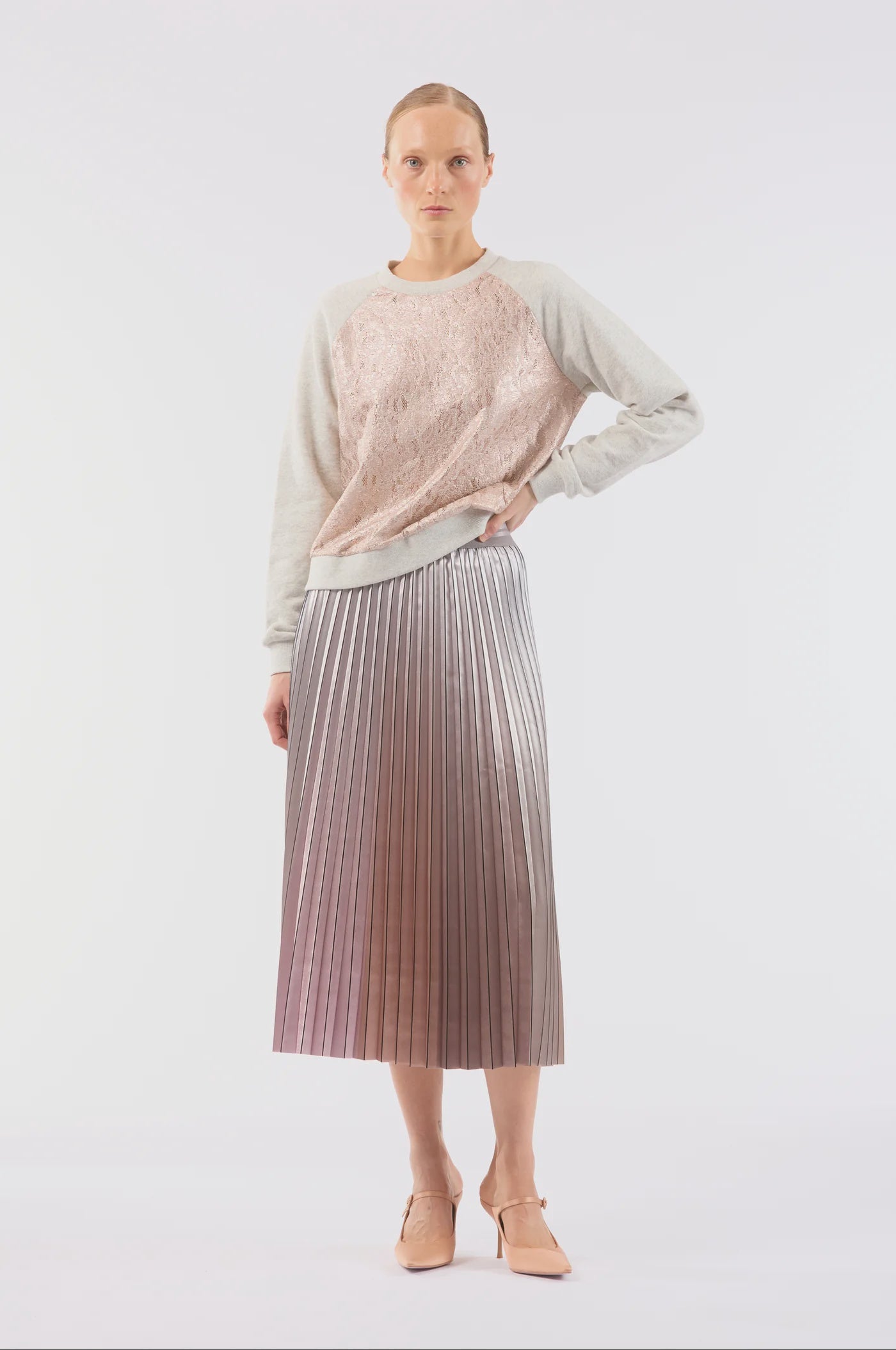 A person poses against a plain white background, dressed in a light pink lace top with long sleeves, the Demure Skirt by Le Superbe—a metallic pleated midi skirt—and light-colored pointed shoes.