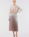 A person poses against a plain white background, dressed in a light pink lace top with long sleeves, the Demure Skirt by Le Superbe—a metallic pleated midi skirt—and light-colored pointed shoes.