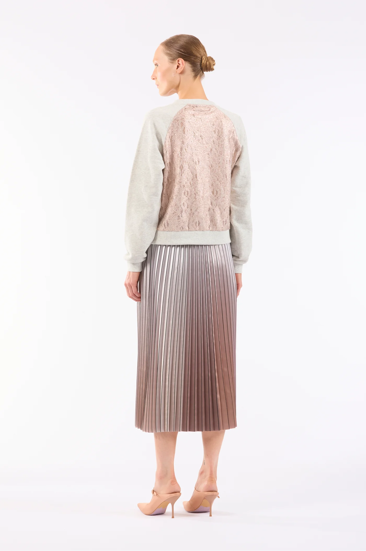 In a white backdrop, someone with a bun hairstyle wears a long-sleeved top with a beige lace back, paired with Le Superbe's stunning metallic degradé midi skirt named Demure Skirt, and nude high heels complete the chic ensemble.