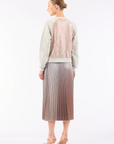 In a white backdrop, someone with a bun hairstyle wears a long-sleeved top with a beige lace back, paired with Le Superbe's stunning metallic degradé midi skirt named Demure Skirt, and nude high heels complete the chic ensemble.