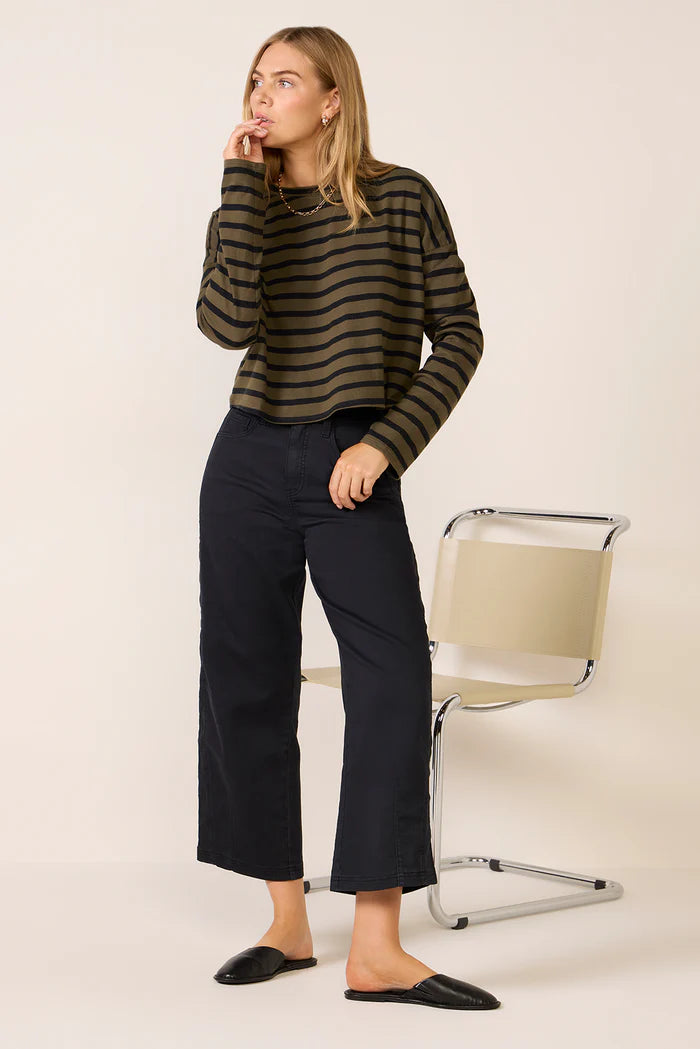 A woman stands indoors wearing the ASKK printed cropped long sleeve shirt, styled with black wide-leg pants and black slip-on shoes. She looks to the side with one hand near her mouth; a modern beige chair is behind her.