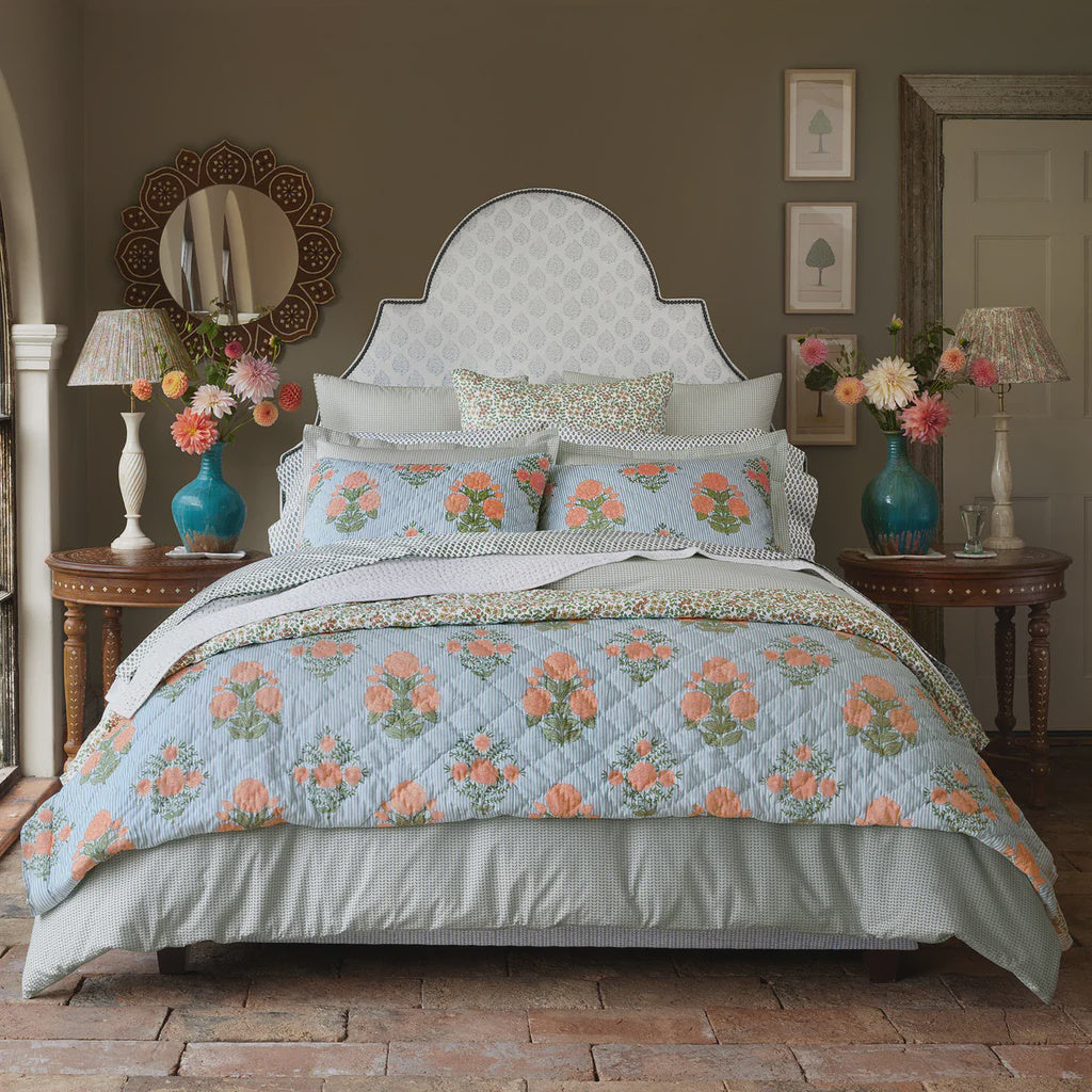 A bed with the John Robshaw Bipin Tangerine Quilt and matching pillows is centered between wooden side tables with turquoise vases. The room has green walls, framed art, and decorative mirrors.