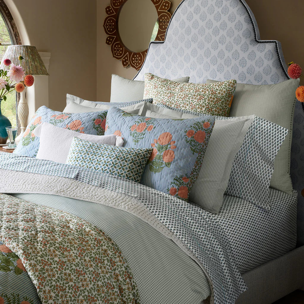 A neatly made bed with layered pillows and bedding in soft blue, green, and white tones showcases the John Robshaw Cala Flat Sheet; a decorative mirror and flower vase sit on a nearby table.