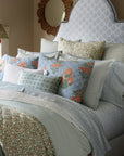 A neatly made bed with layered pillows and bedding in soft blue, green, and white tones showcases the John Robshaw Cala Flat Sheet; a decorative mirror and flower vase sit on a nearby table.