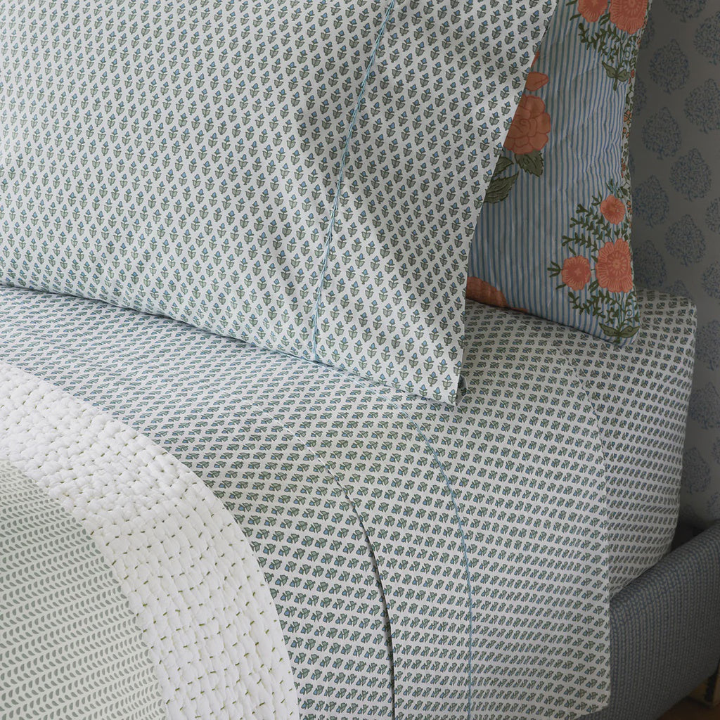 A neatly made bed features the John Robshaw Cala Fitted Sheet in light blue percale with small repeating leaf patterns, striped and floral pillowcases, and a white textured blanket folded at the foot.