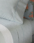 Close-up of a neatly made bed with John Robshaw’s Cala Flat Sheet in light blue, 400 thread count organic cotton, featuring small white floral patterns. One pillowcase shows a blue and white blockprint bouquet with orange flowers. A white textured blanket is folded at the edge.