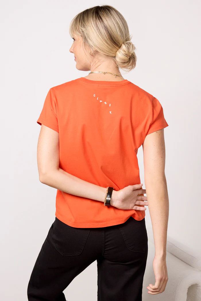 A woman with blonde hair in a low bun wears the Clare Vivier Classic Tee - OUI in orange and black pants, facing away from the camera with her left hand on her back, standing in a minimal indoor space and exuding effortless comfort.