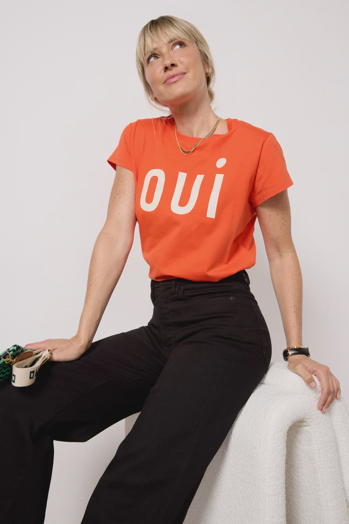 A woman sits on a white chair in comfort, wearing the Clare Vivier Classic Tee OUI Blood Orange and black pants. She gazes upward with a slight smile, holding sunglasses in one hand.